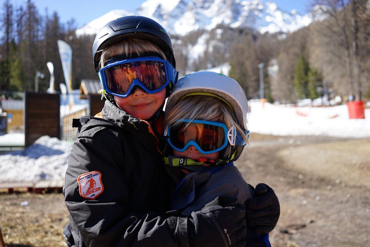 Two children in ski gear and goggles hug each other outdoors with snow and mountains in the background.