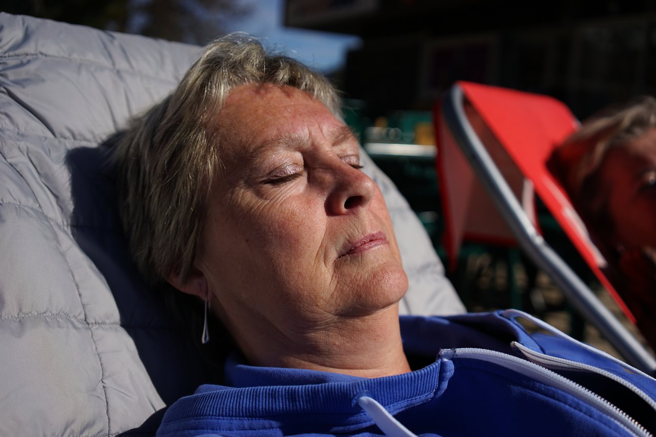 A person in a blue hoodie reclines on a chair with eyes closed, appearing to rest in the sunlight.