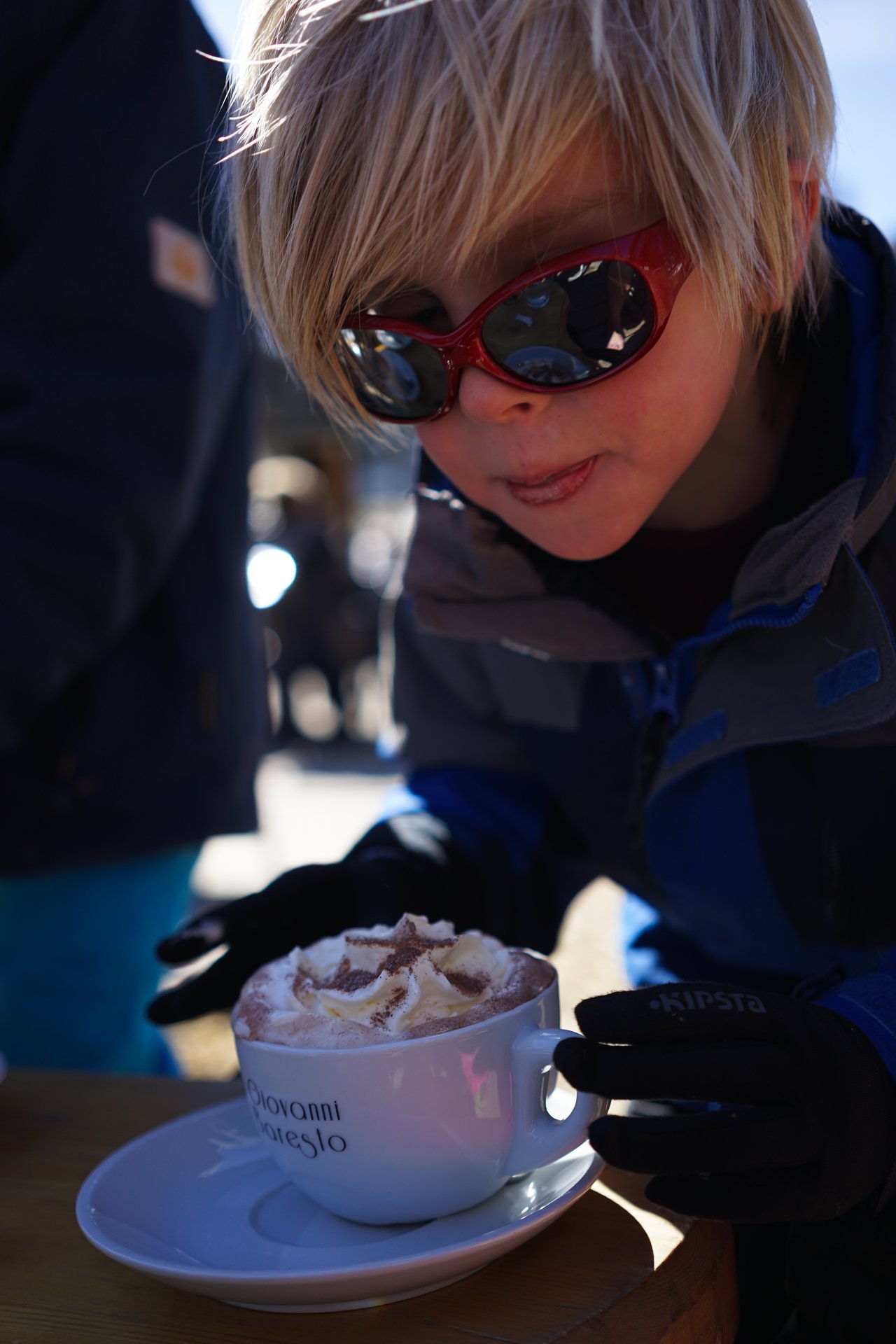 A child wearing sunglasses and gloves leans in to enjoy a cup of hot chocolate with whipped cream.