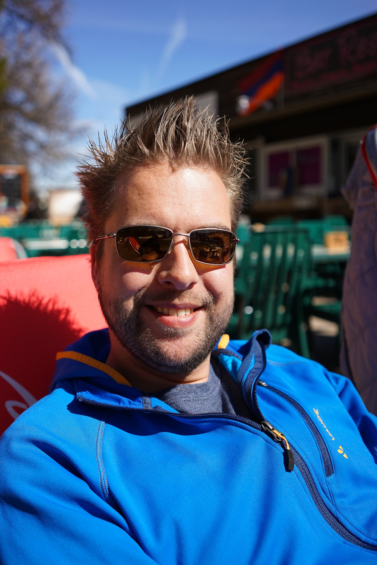 A man wearing sunglasses and a blue jacket smiles while sitting outdoors in the sunlight.