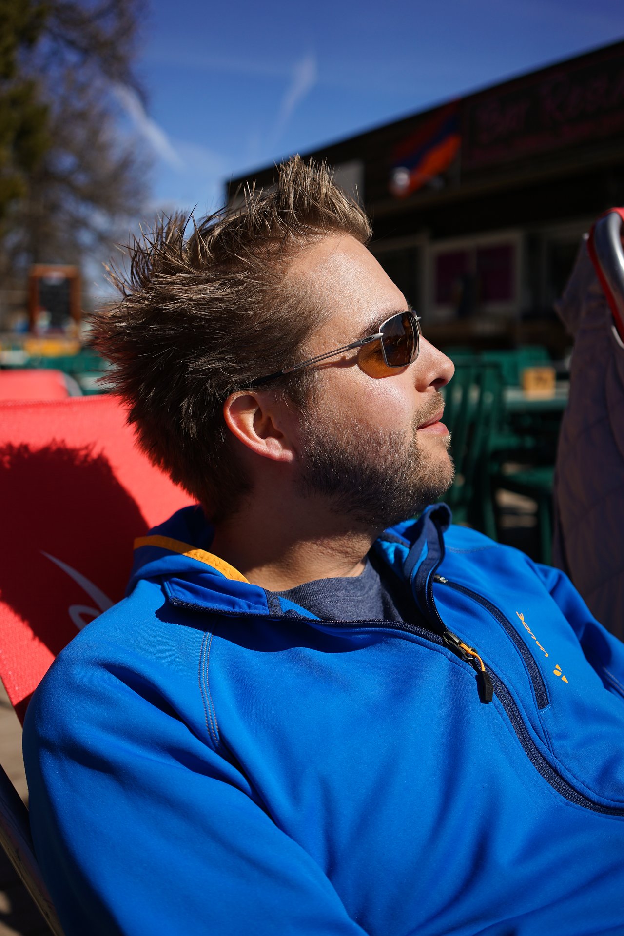 A man in sunglasses and a blue jacket relaxes in a red chair, facing the sunlight.