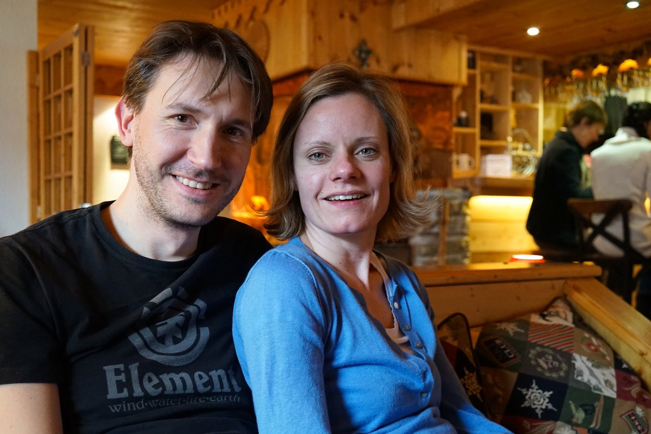 A man and a woman sit closely together on a couch, smiling at the camera in a cozy setting.