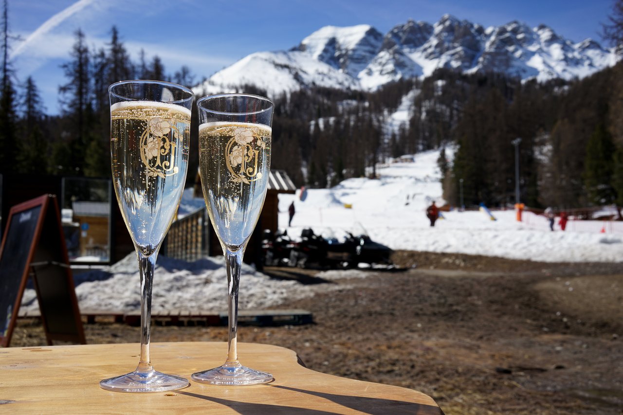 Two glasses of champagne on a wooden table with a snowy mountain and ski area in the background.