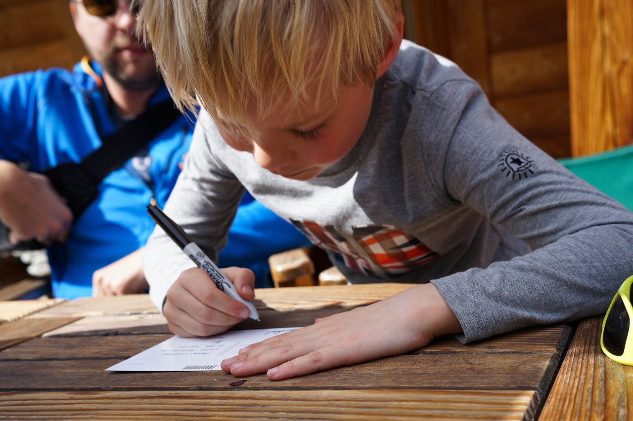 A young child writes on a postcard with a black marker while sitting at a wooden table outdoors.
