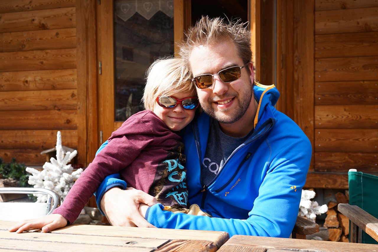 A man in sunglasses hugs a smiling child wearing red glasses while sitting outside a wooden cabin.