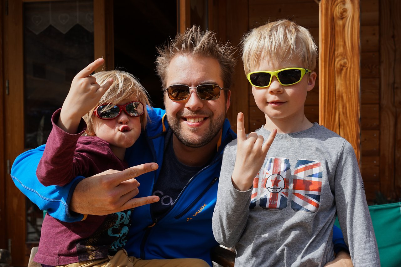 A man and two children wearing sunglasses smile and make rock hand gestures while posing together outdoors.