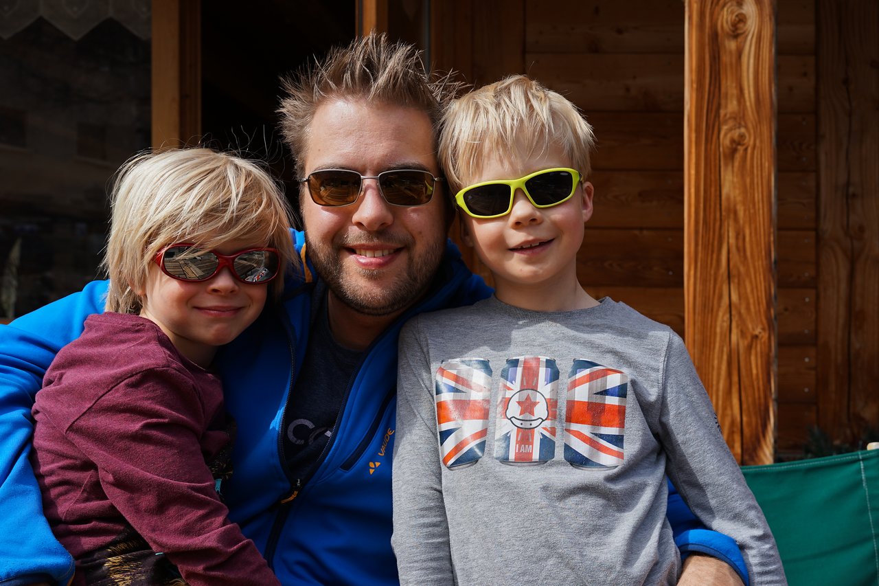 A man in sunglasses hugs two smiling children wearing sunglasses, posing together in front of a wooden building.