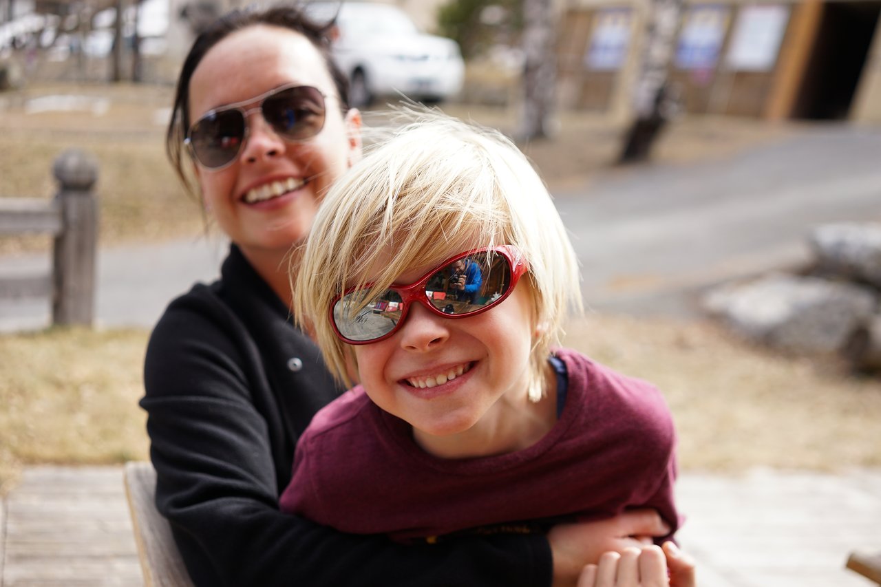 A smiling child wearing red sunglasses leans forward while an adult behind them hugs them from behind.