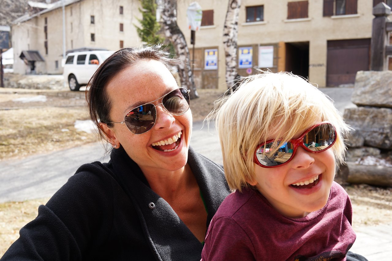 A woman and a child wearing sunglasses smile while sitting outdoors in a sunny setting.