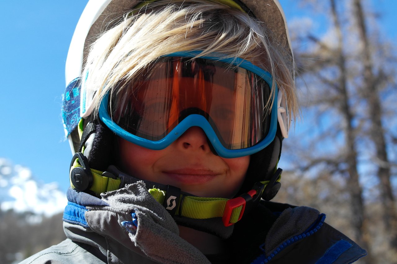 A child wearing ski goggles and a helmet smiles while dressed in winter gear outdoors.