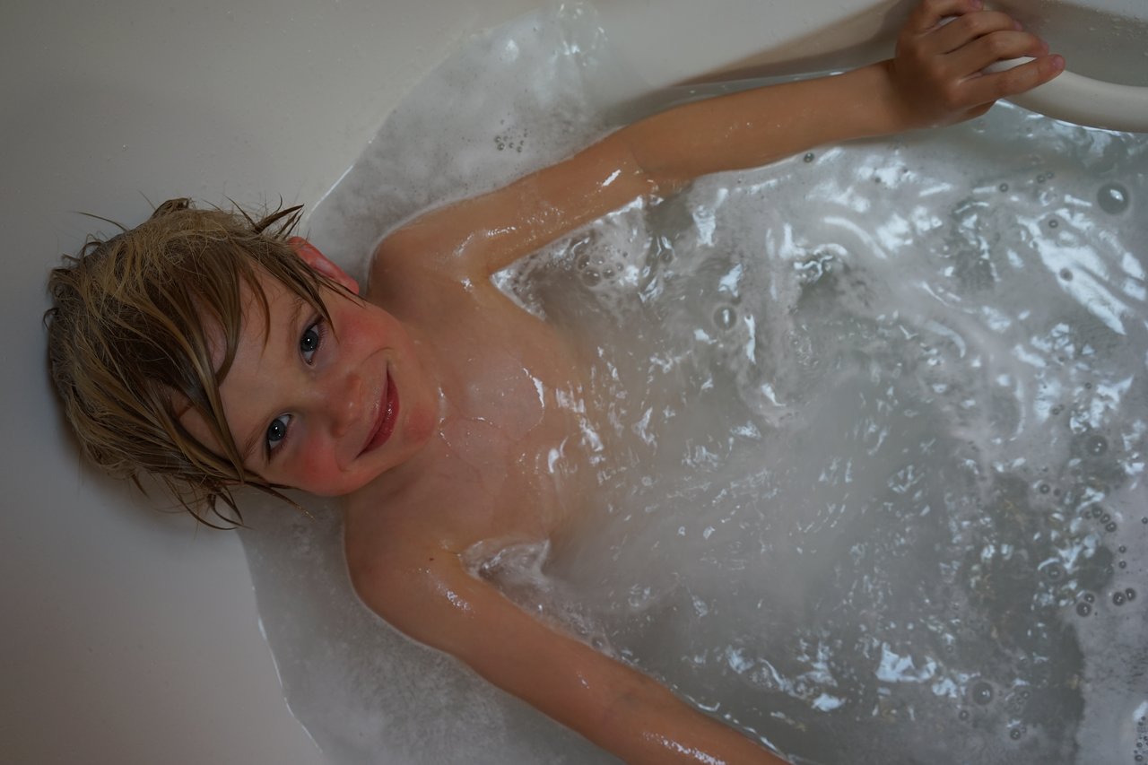 A child with wet hair smiles while relaxing in a bathtub filled with water and bubbles.
