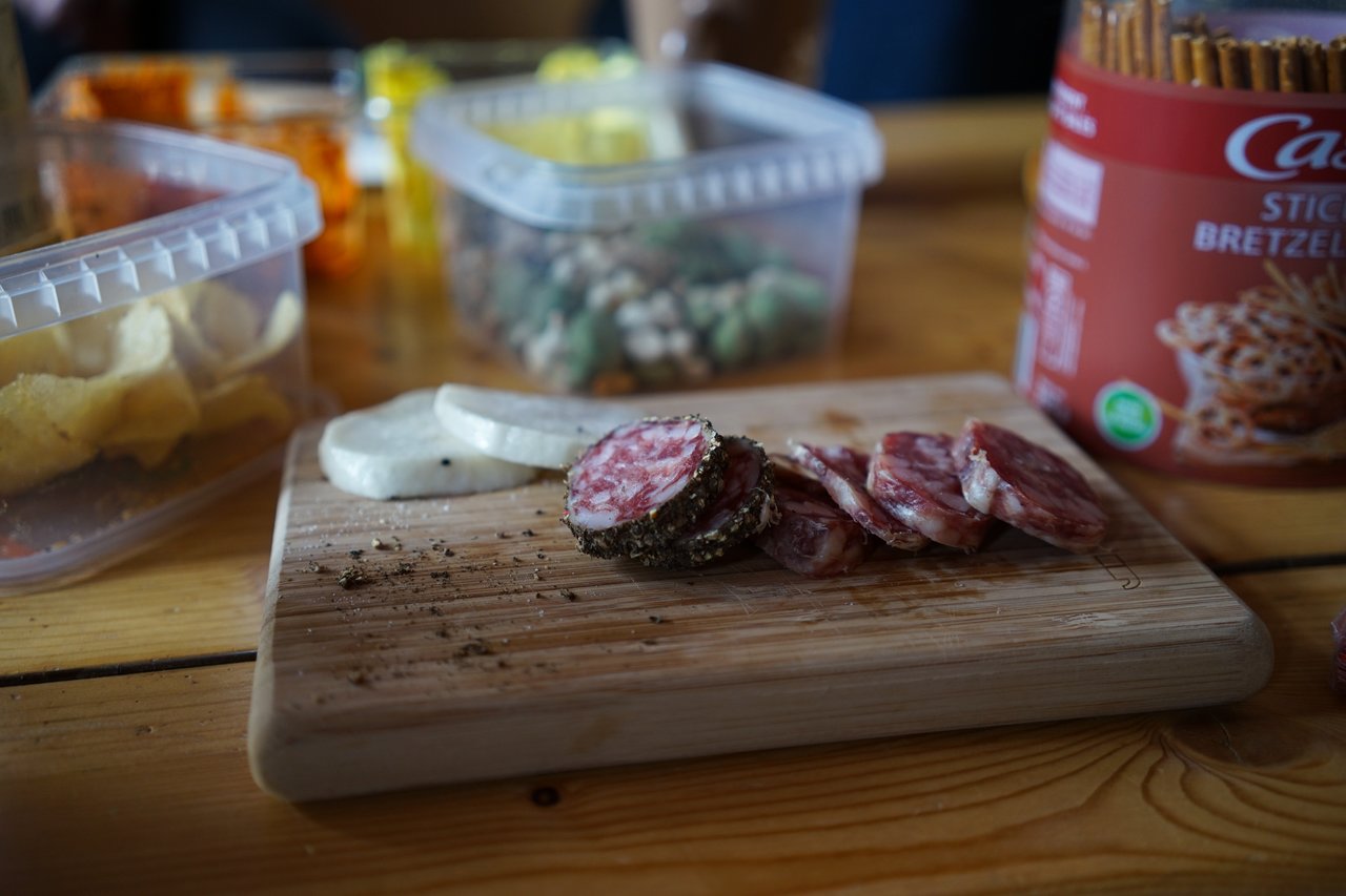 Sliced sausage and cheese on a wooden cutting board, with snacks in plastic containers on a table.