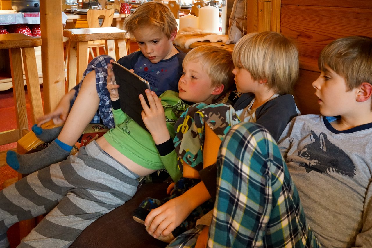 Four children in pajamas sit closely together on a couch, watching a tablet screen with focused expressions.