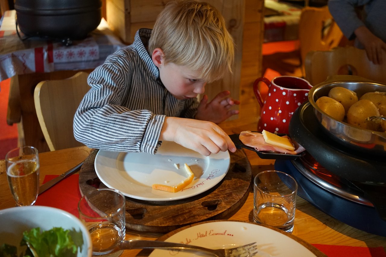 A child in a striped shirt carefully places cheese on a plate while preparing a meal at the table.
