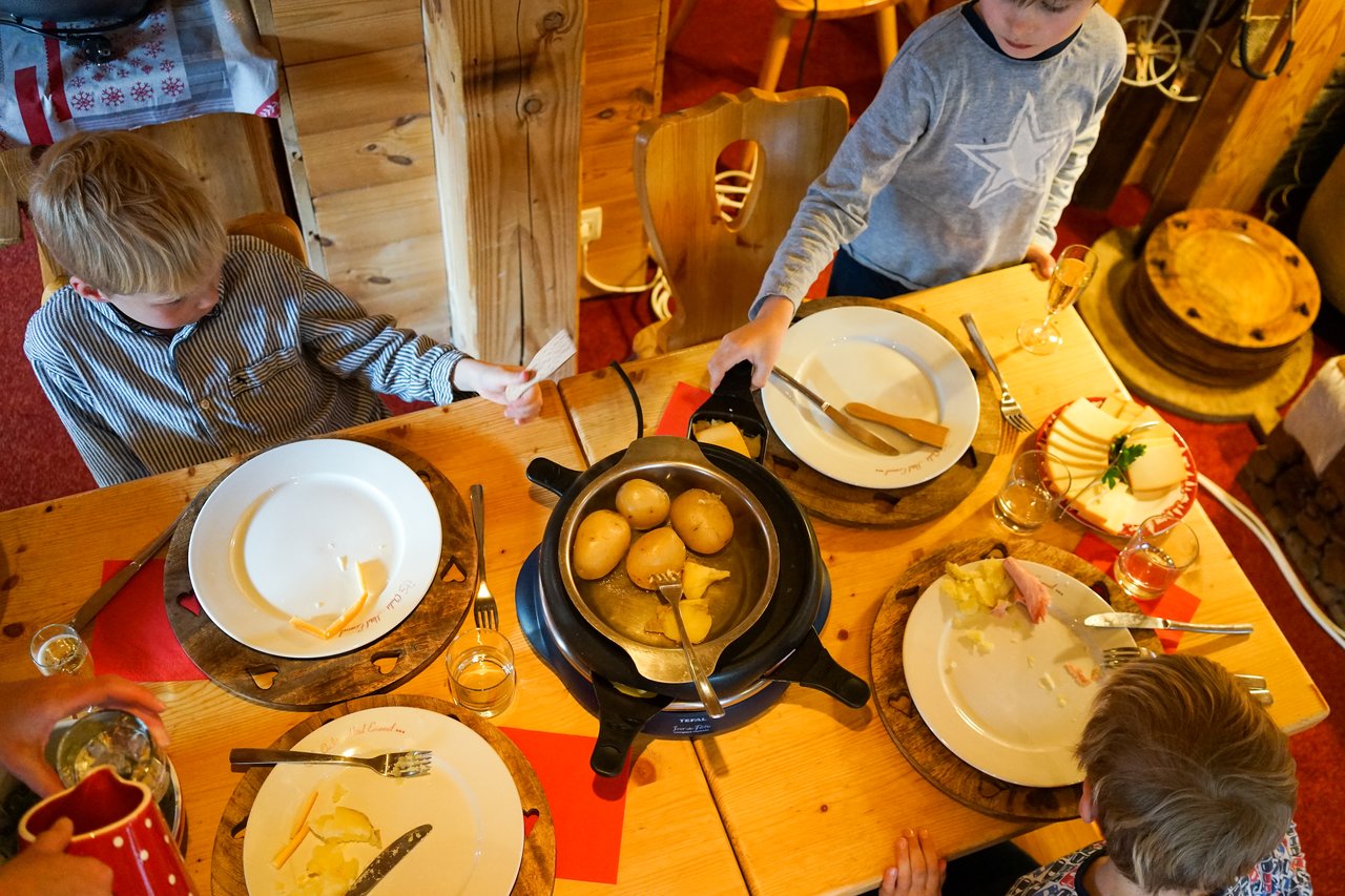 Children sit around a wooden table with plates, cheese, and a pot of boiled potatoes in the center.
