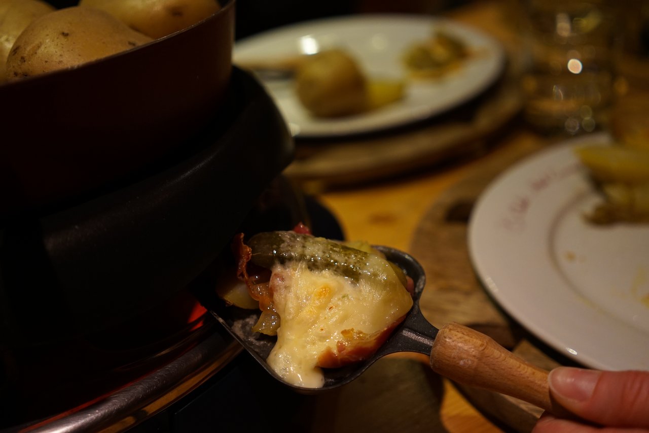 A person holds a small pan with melted cheese, pickles, and ham, preparing a raclette meal.