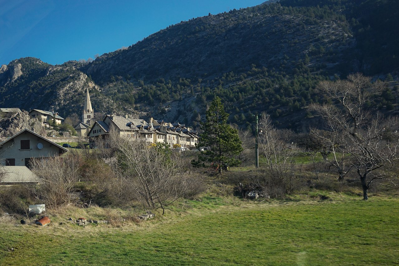 A small village with stone houses and a church steeple, surrounded by green hills and mountains.