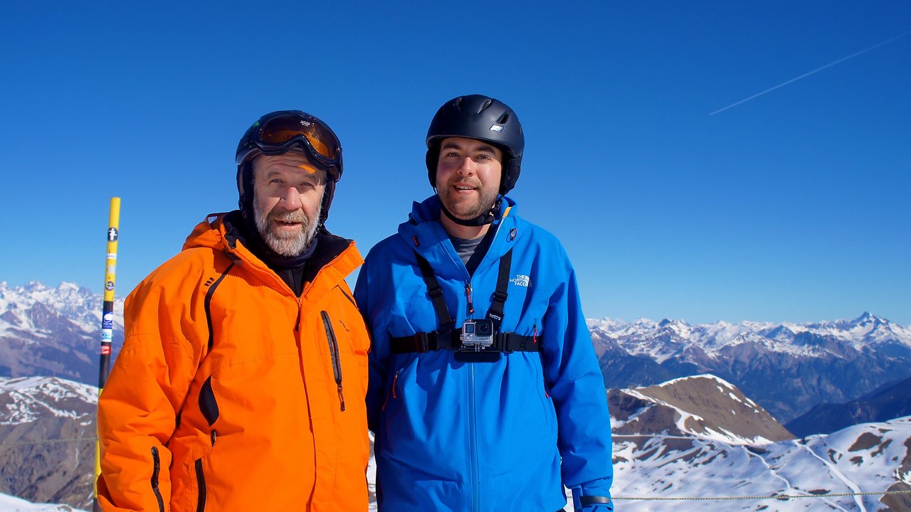 Two people in ski gear stand on a snowy mountain, smiling at the camera.