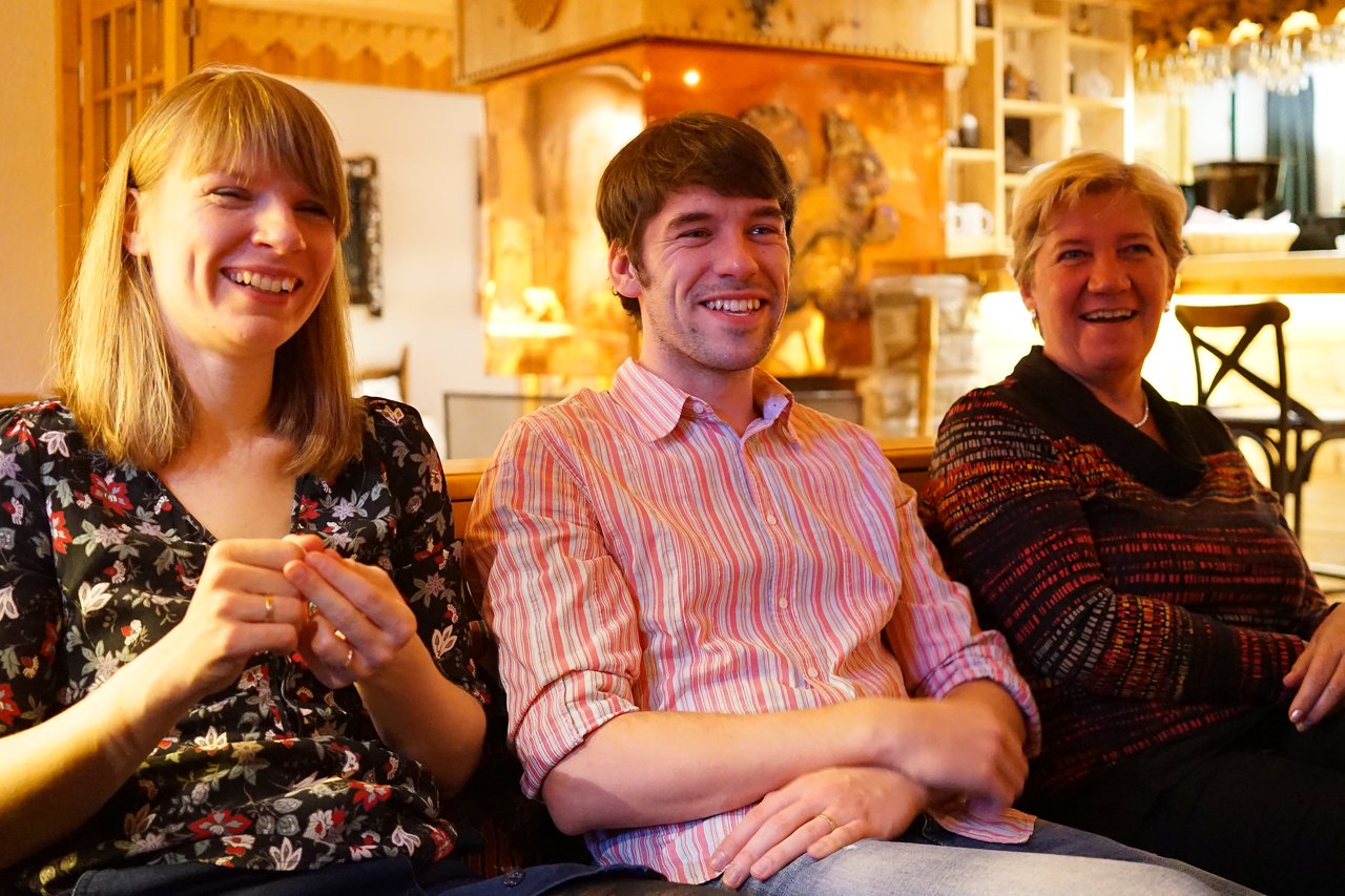 Three people sitting indoors, smiling and laughing while looking at something off-camera.
