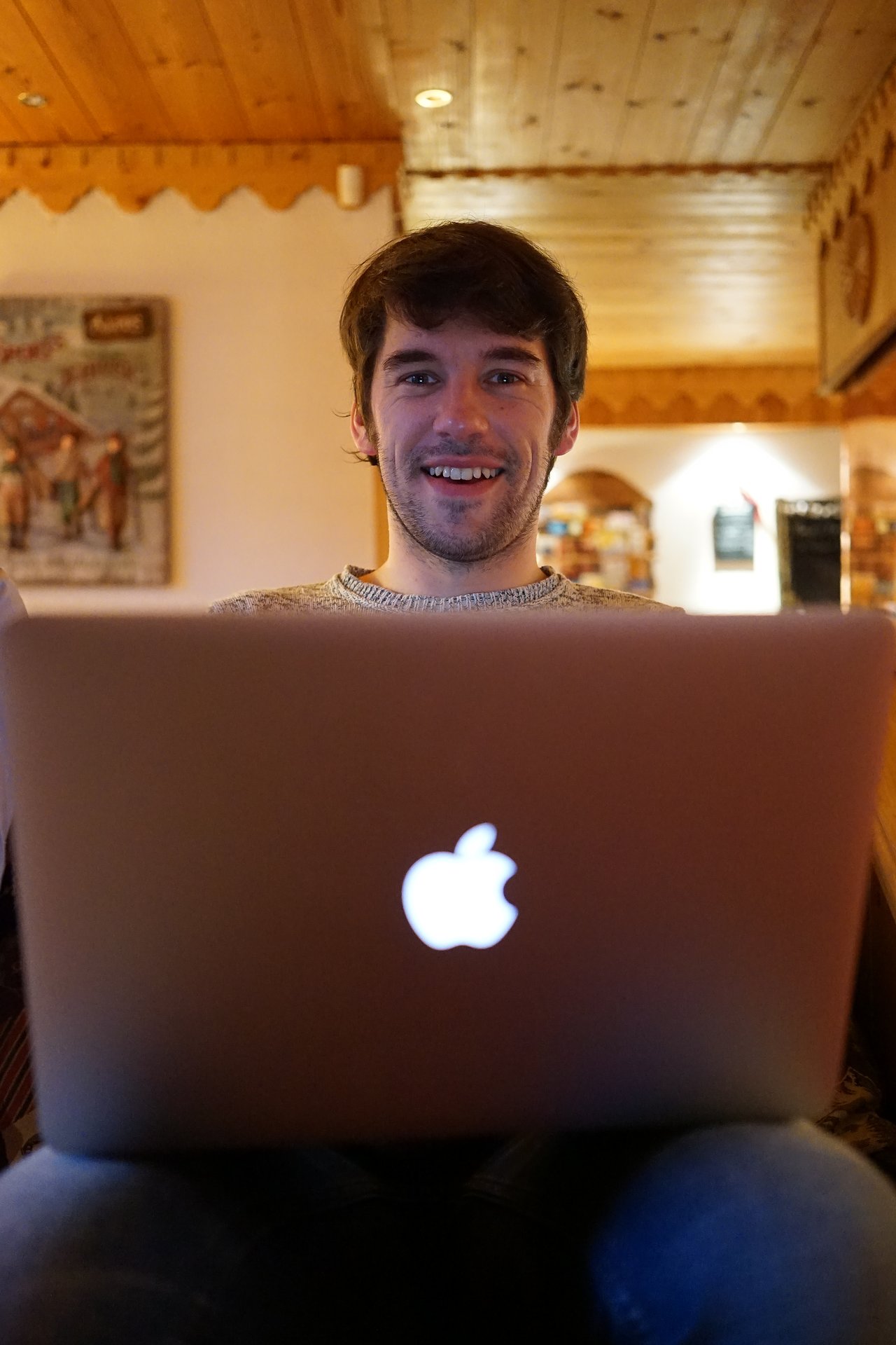 A smiling person sits indoors, looking at a laptop with an illuminated Apple logo on the back.