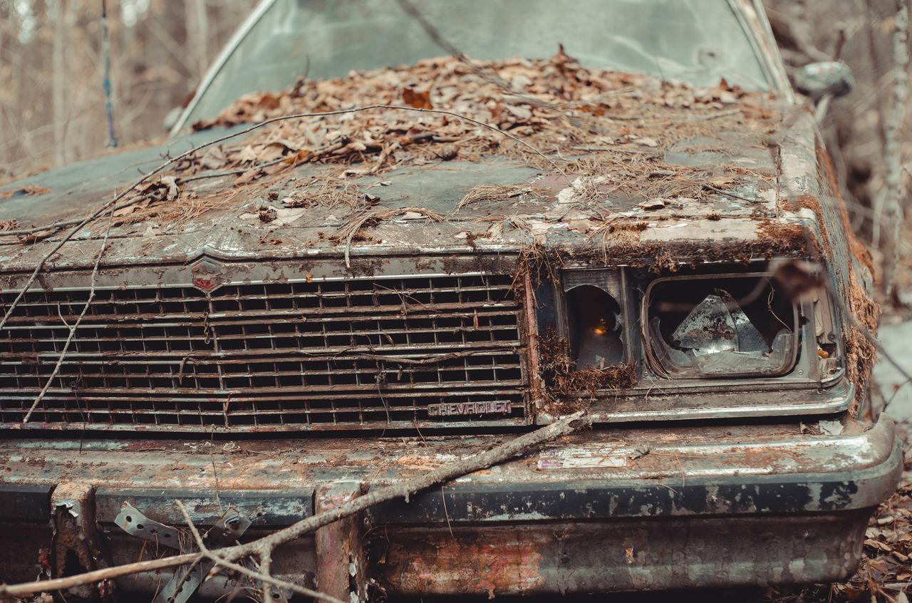 An old, abandoned car covered in leaves and dirt sits in the woods with broken headlights and rusted parts.