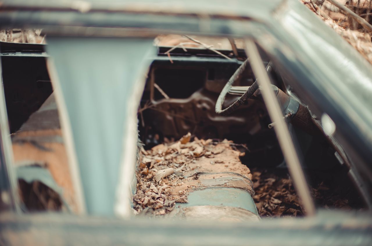 Rusty, abandoned car in the woods with broken windows and a decayed interior covered in dirt and fallen leaves.