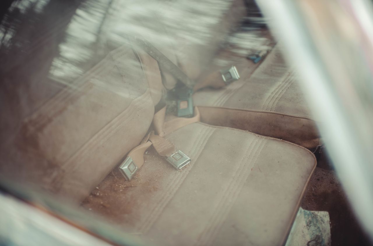 Close-up view through a car window showing worn beige seats and old seat belts inside an abandoned vehicle.
