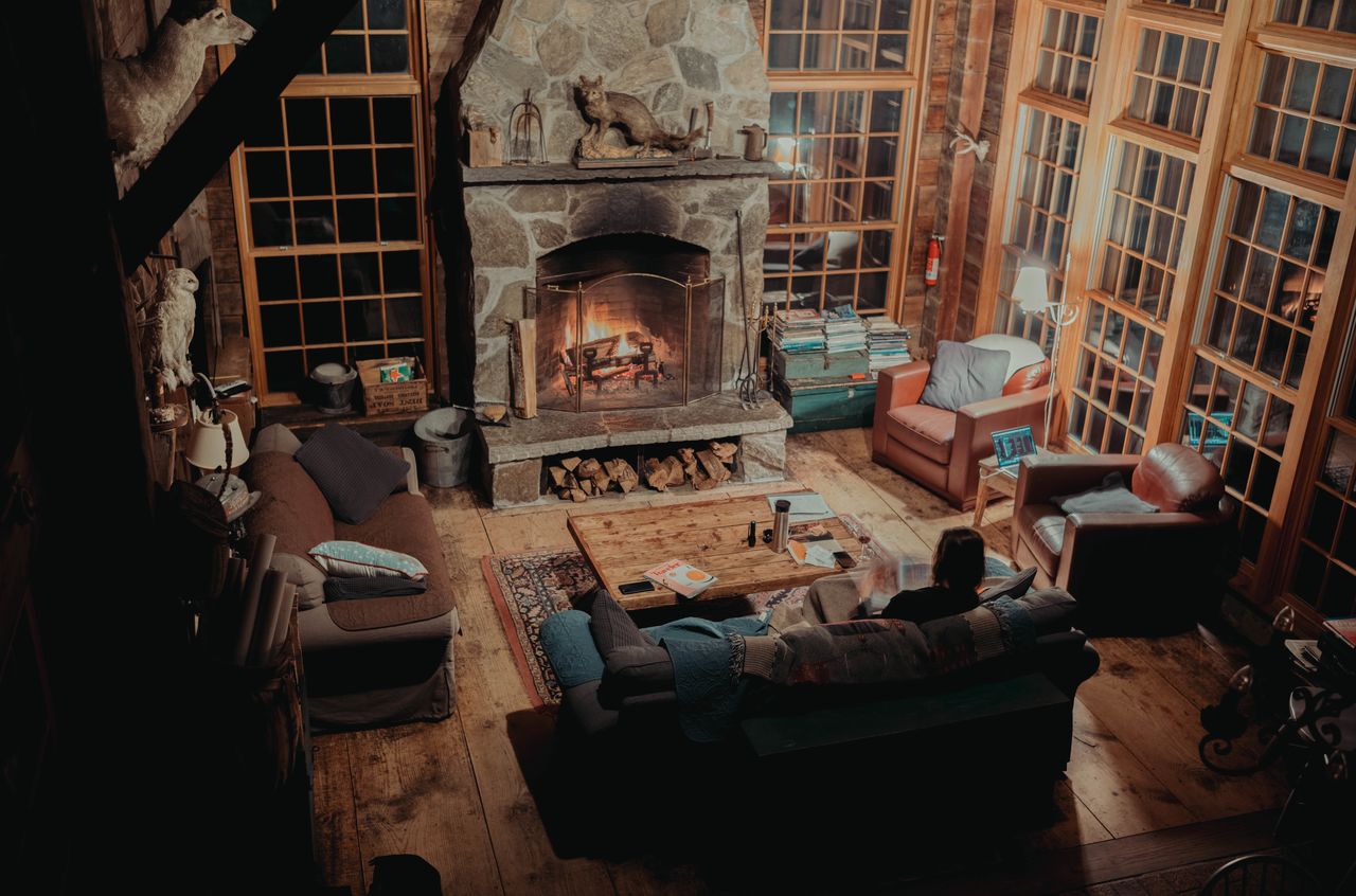 A person sits on a couch by a fireplace, reading and relaxing in a cozy cabin at night.
