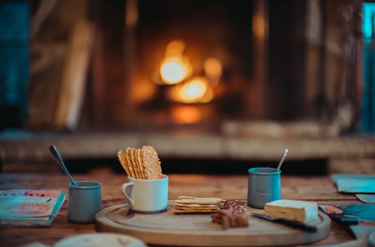 Crackers, cheese, and hot drinks on a wooden table in front of a warm fireplace at night.