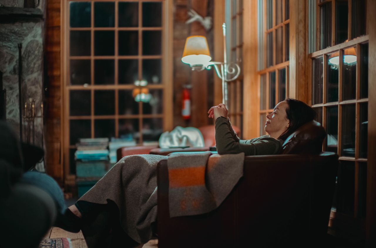 A person reclines in a cozy chair at night, covered with a blanket, appearing relaxed and content.