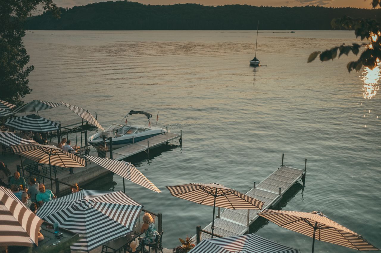 Lakeside restaurant with docked boats, people dining under umbrellas, and a sunset.