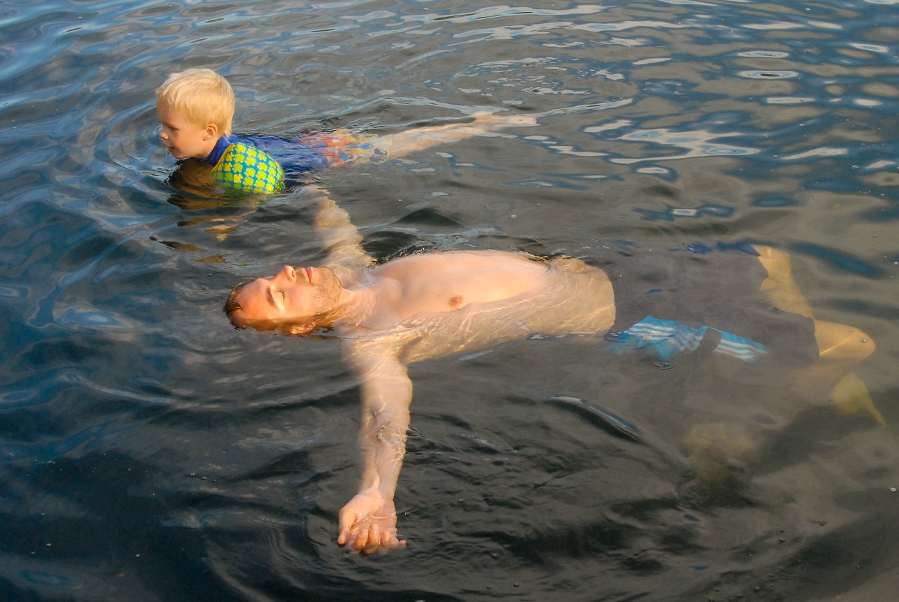 A child and an adult swim in a quarry, with the child paddling and the adult floating on their back.