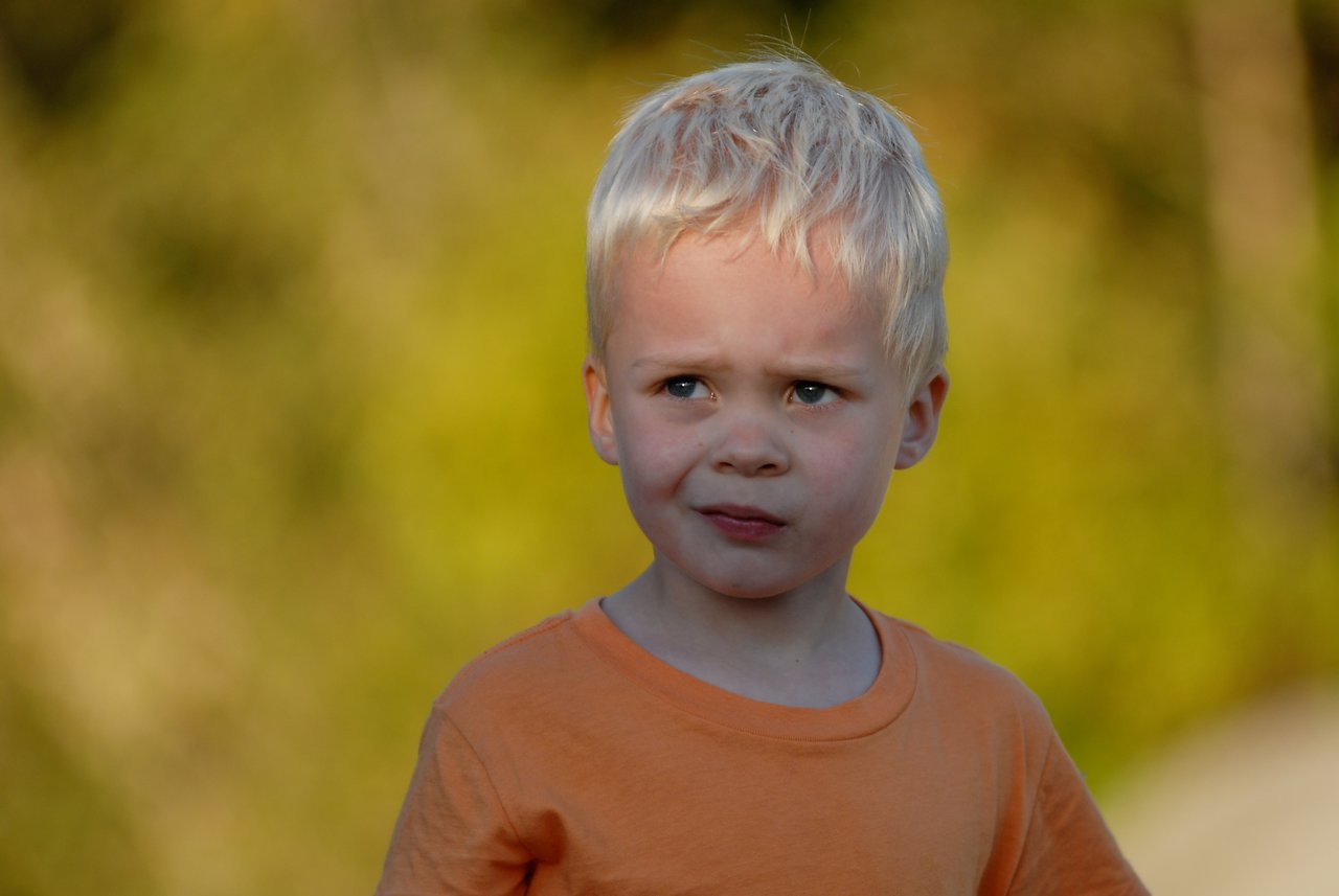 A young child with blond hair and an orange shirt looks concerned while gazing at something off-camera.