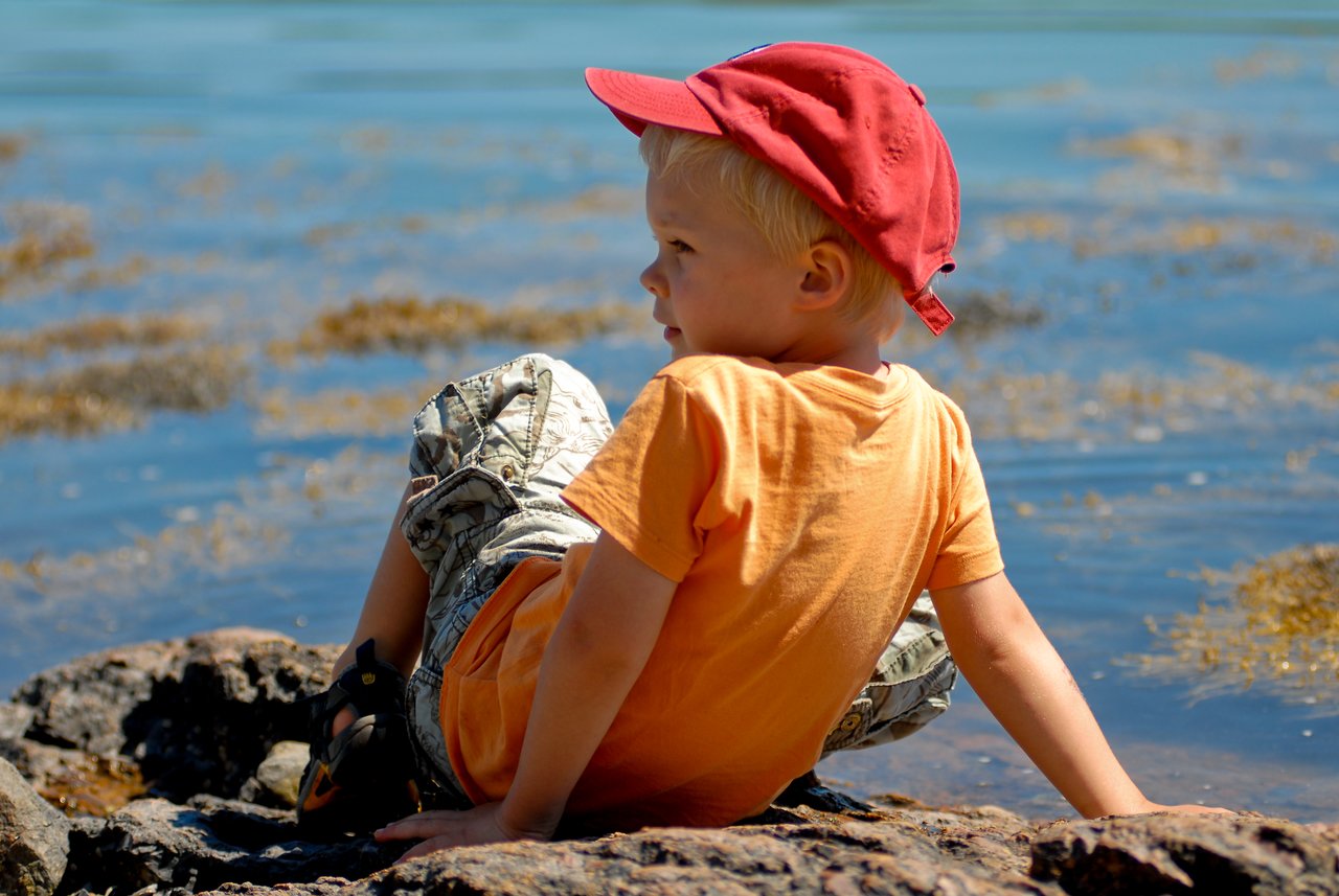 A young child in an orange shirt and red cap sits on a rock near the water, looking away.