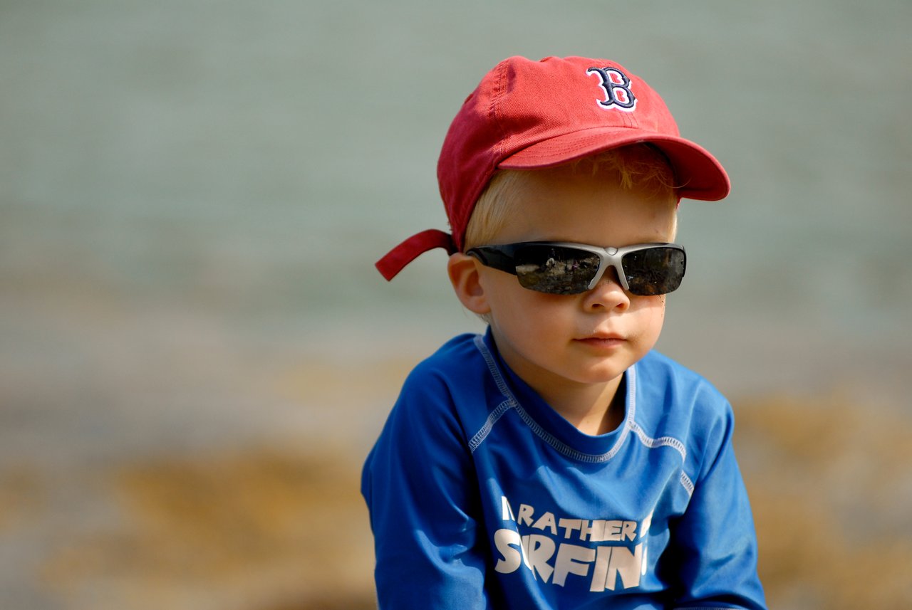 A young child wearing sunglasses, a red baseball cap, and a blue shirt sits outdoors with a neutral expression.
