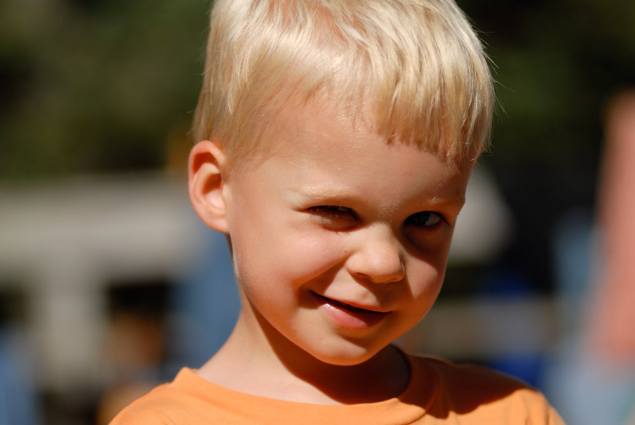 A young child with blonde hair and an orange shirt winks playfully at the camera.
