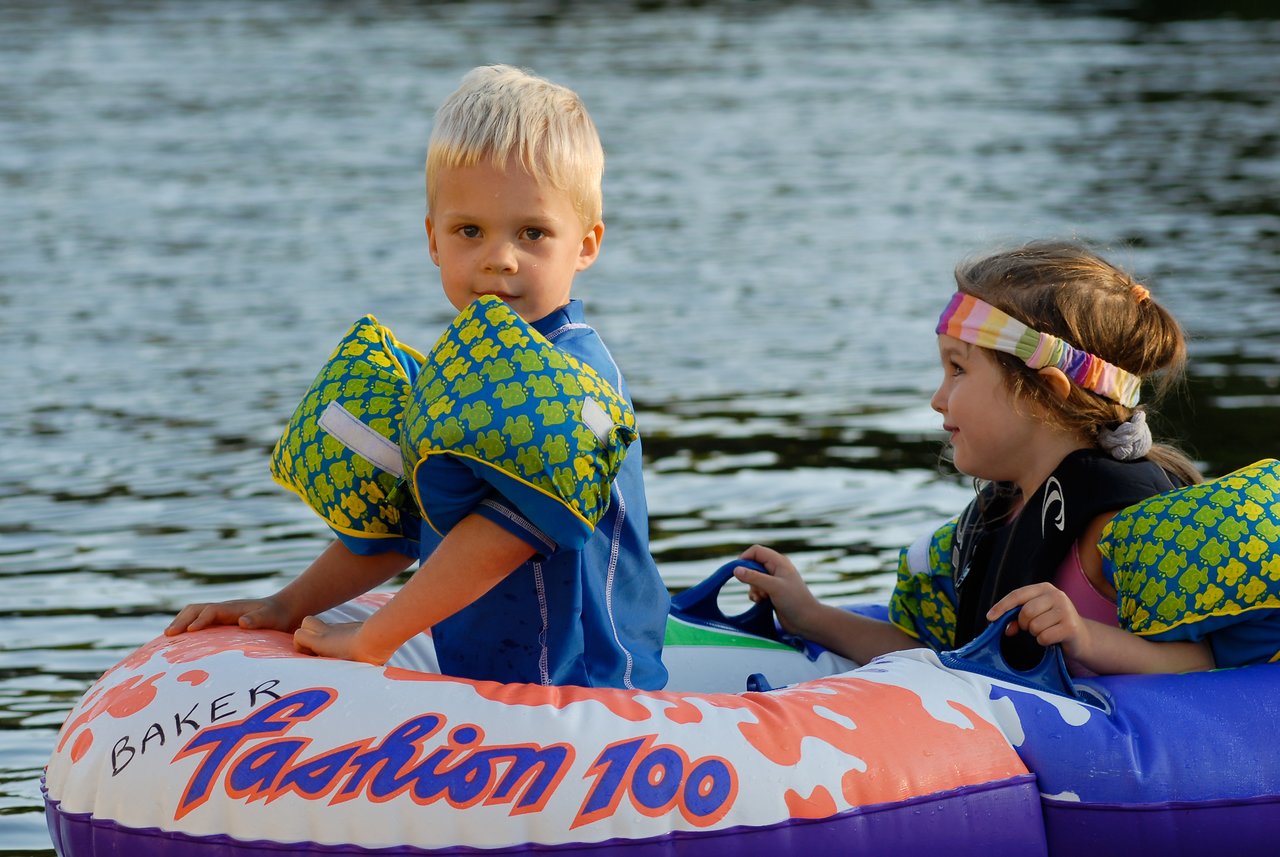 Two young children wearing floatation gear sit in an inflatable boat on the water, holding onto the handles.