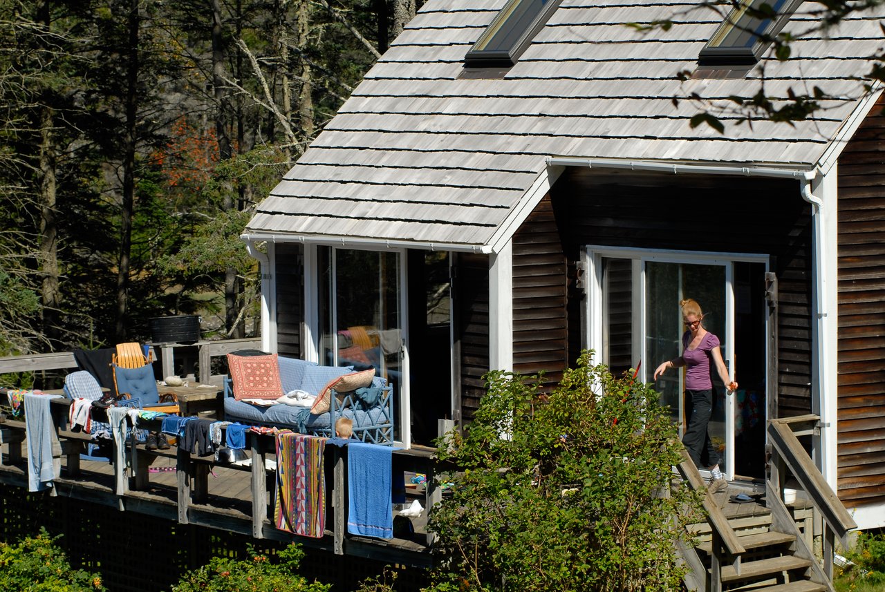 A person steps out of a cabin onto a wooden deck with laundry hanging and someone lounging on a chair.