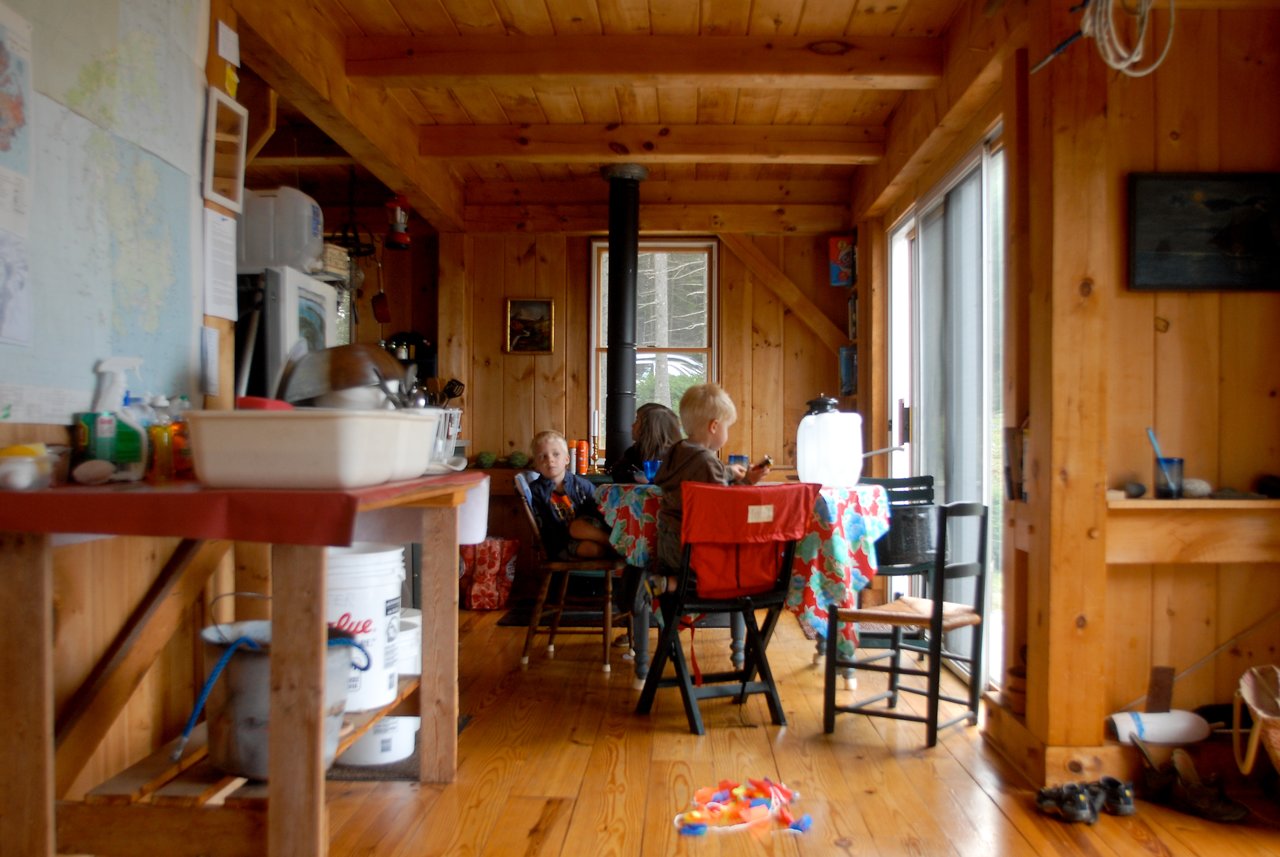 Three children sit at a wooden table inside a cabin, engaged in an activity.