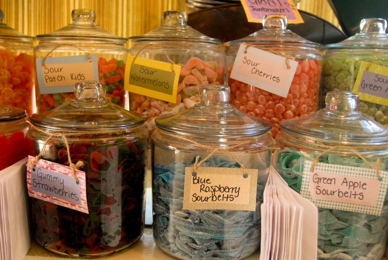Glass jars filled with colorful candies, including gummy strawberries, sour cherries, and sourbelts, labeled with handwritten tags.