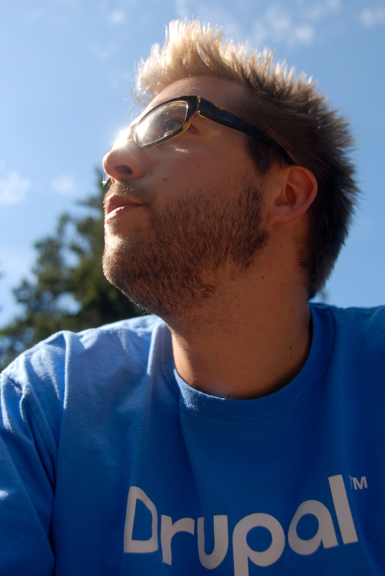 A man wearing glasses and a blue Drupal T-shirt looks up with sunlight on his face.