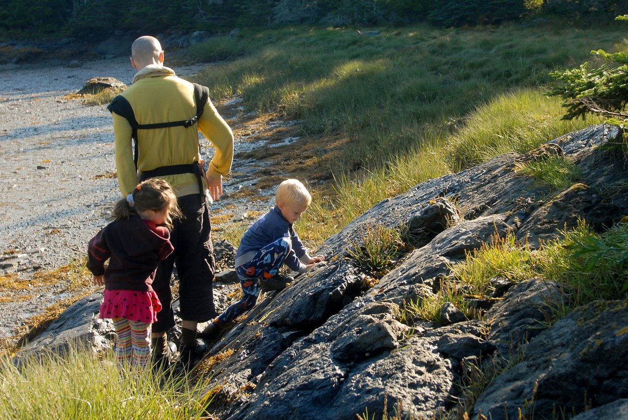 An adult and two children explore a rocky, grassy area near a beach, with one child climbing a rock.