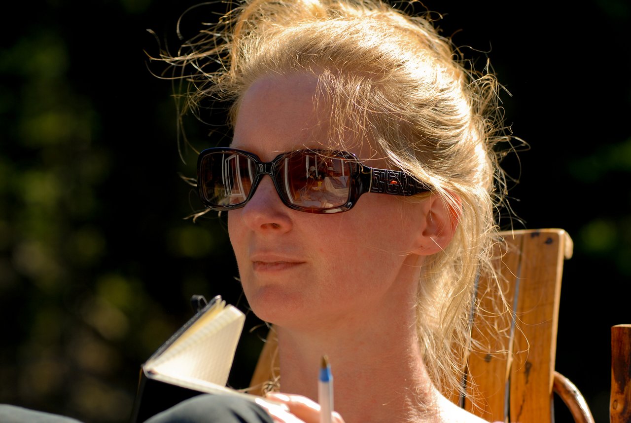 A person wearing sunglasses sits outdoors, holding a pen and notebook, appearing to be in thought.