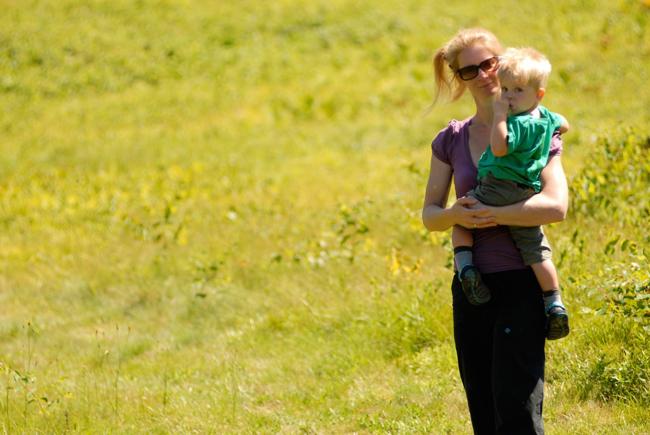 A woman wearing sunglasses holds a young child in her arms while standing in a grassy field.