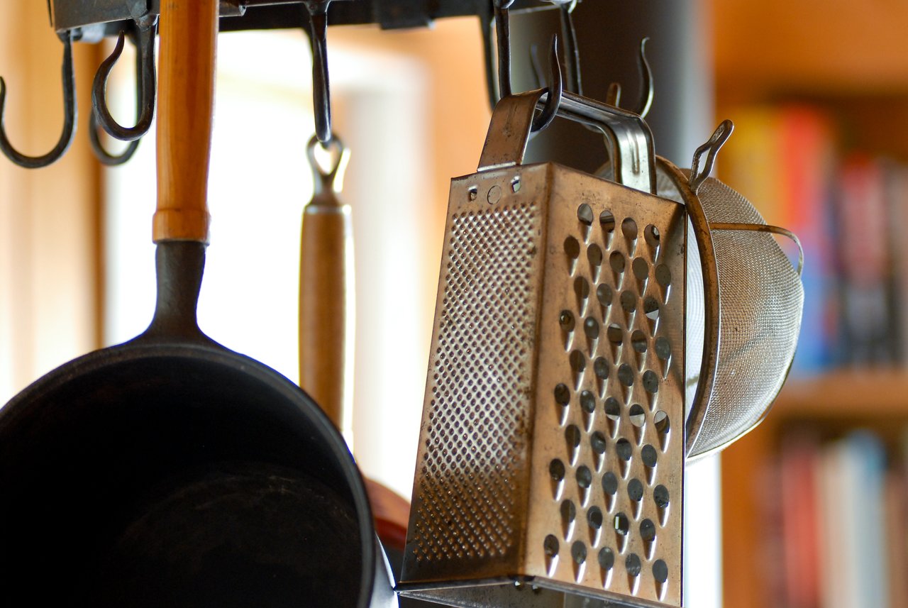 Metal kitchen utensils, including a grater, strainer, and frying pan, hang from hooks in a well-lit space.