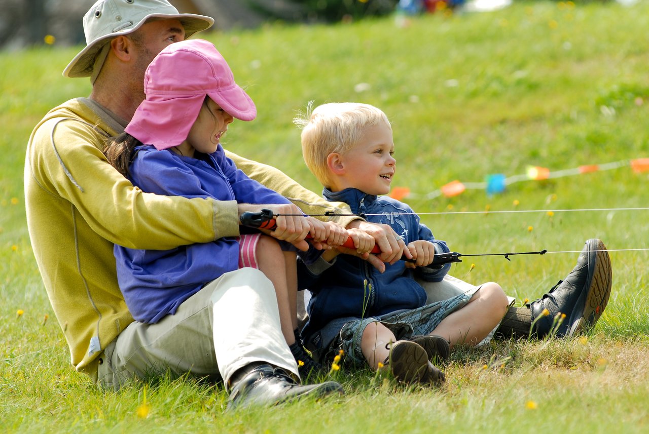 An adult and two children sit on the grass, holding kite handles and smiling while flying a kite together.