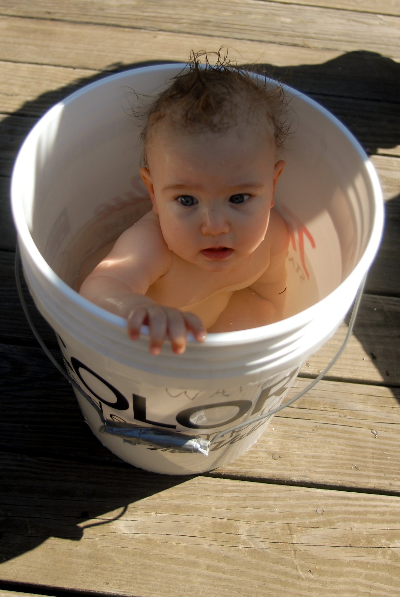 A baby with wet hair sits in a large plastic bucket, looking up while gripping the edge.
