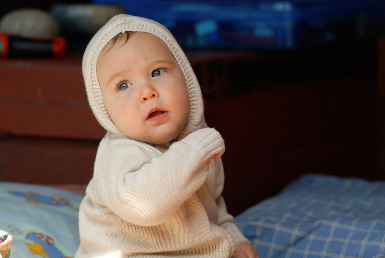 A baby in a beige hooded sweater sits on a cushion, looking up with a curious expression.