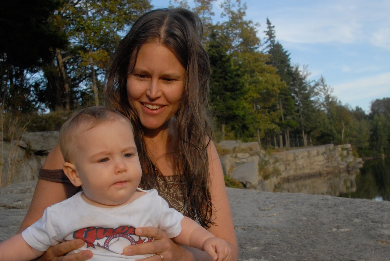 A woman sits outdoors, smiling while holding a baby who looks off to the side.