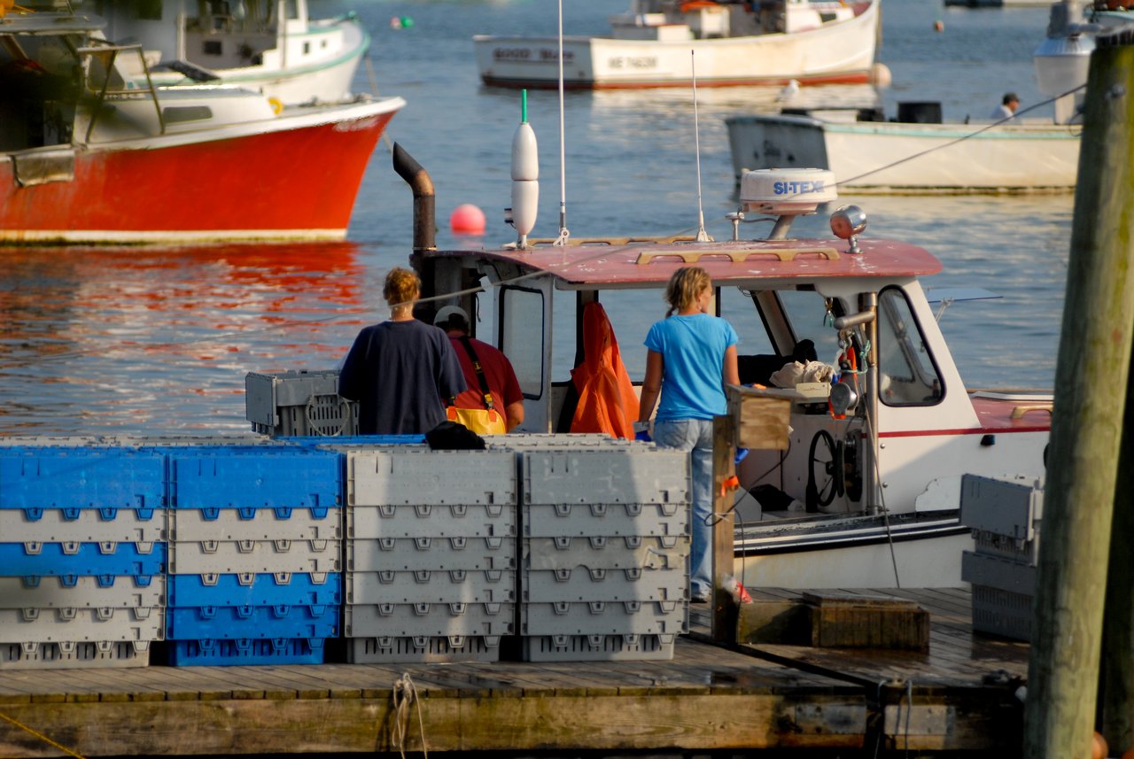 Two people stand on a dock next to a lobster boat, handling crates filled with seafood.